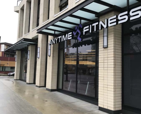 Street view of an Anytime Fitness gym with large windows and a glass awning, located in a modern beige brick building on a wet sidewalk.
