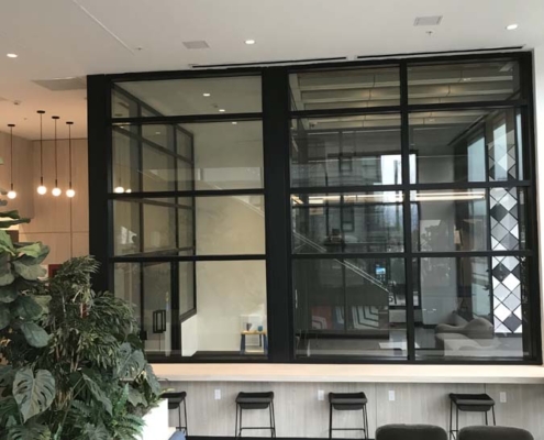 A modern office lounge with green plants, blue seating, black armchairs, and a high counter with stools; large glass windows reveal a bright stairwell and more seating in the background.