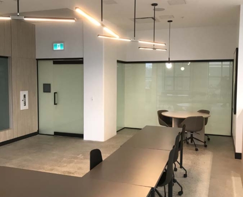 Modern office space with black chairs around long black tables, minimalist decor, pendant ceiling lights, glass doors, and light-colored walls and flooring. The area appears clean and empty, with natural light coming in.