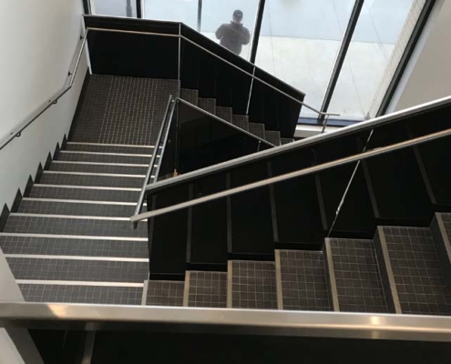 A view looking down a modern indoor staircase with black steps and metal railings, leading to large floor-to-ceiling windows. Outside, a person is visible on the sidewalk. Several ceiling lights hang above.