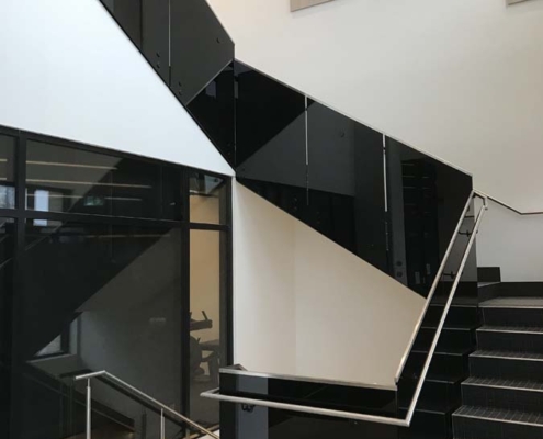 A modern indoor staircase with black glossy panels and stainless steel handrails, set against white walls and glass partitions in a well-lit building interior.