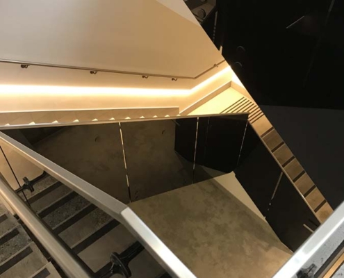 A modern indoor staircase with metal railings and glass panels, illuminated by warm, recessed lighting along the walls and steps, creating geometric reflections on the surfaces.