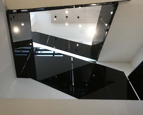 View looking up through a modern, geometric staircase with glossy black railings and white walls, illuminated by round ceiling lights. The stair landings create angular shapes.