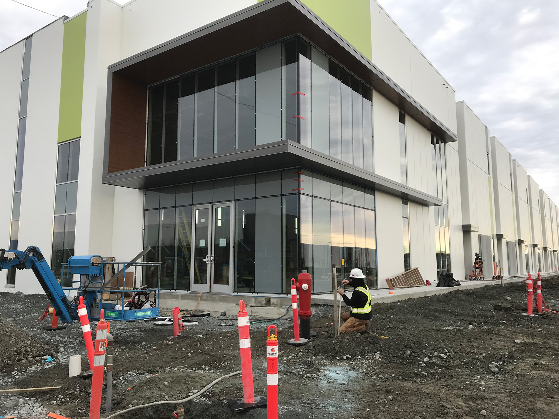 A construction worker kneels by a modern building with large glass windows and white walls. The ground is unfinished, with construction cones, equipment, and dirt surrounding the site.