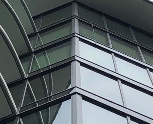 Close-up view of the corner of a modern building with large glass windows and metal frames, featuring curved architectural details and an overhanging roof against a cloudy sky.