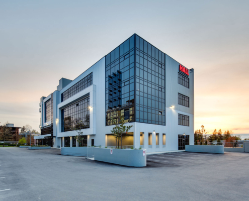 A modern, three-story office building with large glass windows and the red HAO logo on the top corner, set against a sunset sky with an empty parking lot in front.