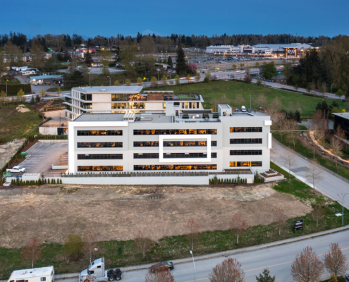 A modern four-story office building with large windows is surrounded by greenery and parking lots. Other buildings, roads, and trees are visible in the background during twilight.