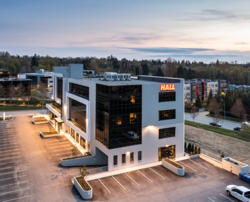 A modern, four-story office building with large glass windows and a HALL sign illuminated on the corner, surrounded by an empty parking lot and trees at dusk.