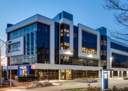 A modern, multi-story office building with large glass windows and geometric design, illuminated at dusk. The parking lot in front is mostly empty, and a few trees border the property.