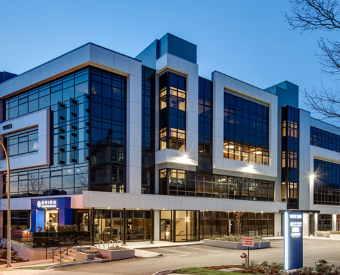 A modern, multi-story office building with large glass windows and geometric design, illuminated at dusk. The parking lot in front is mostly empty, and a few trees border the property.
