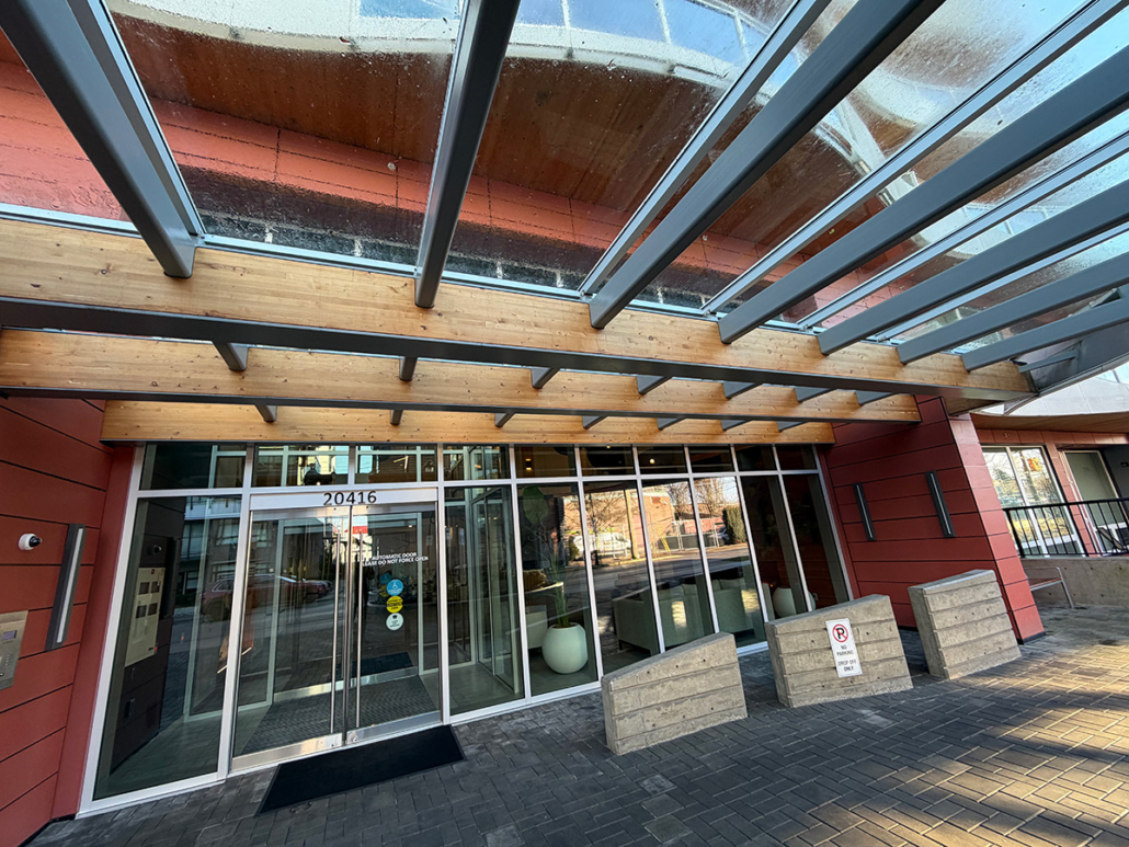 Entrance to a modern building with large glass doors and windows, a wooden and metal awning overhead, and a paved walkway leading up to the door. Reflections and outdoor scenery are visible through the glass.