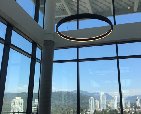 A modern room with large floor-to-ceiling windows overlooks a cityscape with tall buildings, green trees, and distant mountains under a clear blue sky. A circular ceiling light hangs in the foreground.