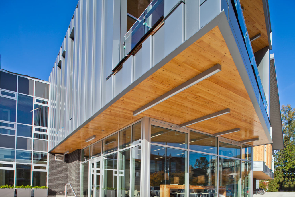 Modern building exterior with large glass windows, metal siding, and a wooden overhang. The angular architecture features clean lines and an open, contemporary design, with sunlight illuminating the entrance area.