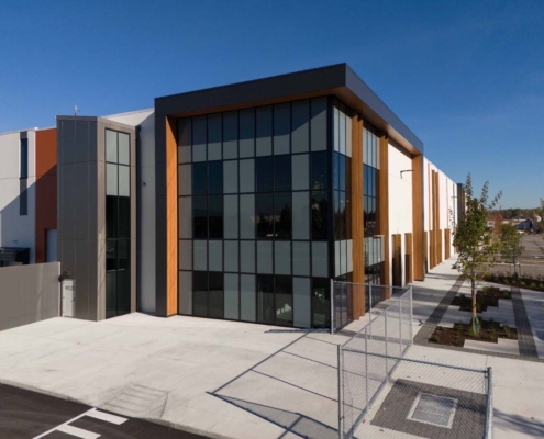 A modern, two-story commercial building with large glass windows, wood panel accents, and a clean, paved exterior surrounded by young trees and minimal landscaping under a clear blue sky.