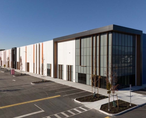 A modern, large warehouse or industrial building with glass and panel facades sits in an empty parking lot under a clear blue sky. Young trees are planted along the sidewalk.