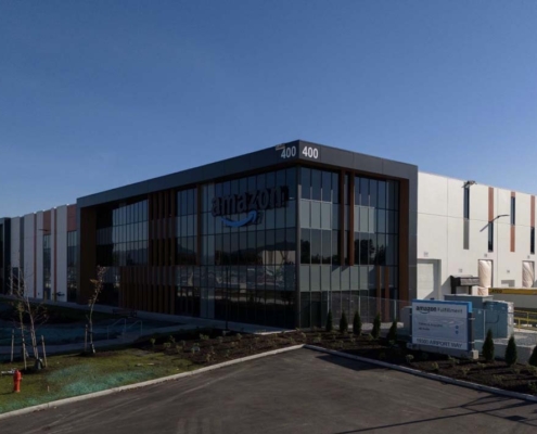 A large Amazon warehouse with a modern glass facade, labeled “400,” sits under a clear blue sky. The building has the Amazon logo and is surrounded by landscaping and a parking area.