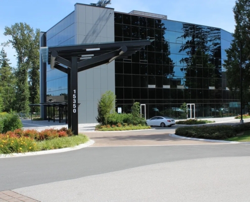 A modern glass office building with the number 15350 displayed on a black signpost near the entrance, surrounded by greenery and a parked silver car on a sunny day.