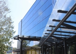 A modern glass office building with reflective blue windows and a black metal and glass awning, surrounded by trees and a clear sky.