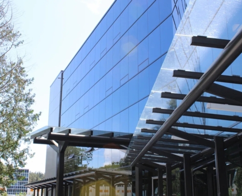 A modern glass office building with reflective blue windows and a black metal and glass awning, surrounded by trees and a clear sky.