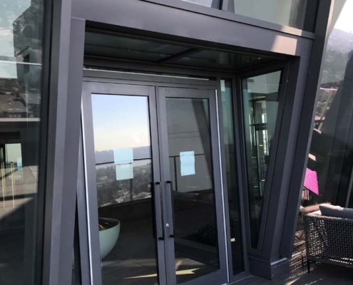 Glass double doors on a modern building with angled metal frames; two papers are taped to the doors, and a city skyline is visible in the reflection.