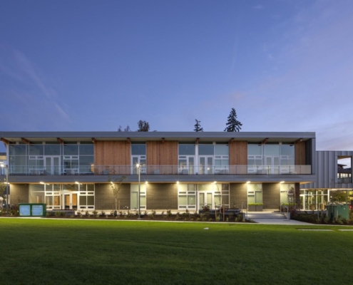 A modern two-story building with large windows and wood panel accents is seen at dusk, with lights glowing inside. The building faces an open, grassy lawn and is surrounded by landscaping and trees.