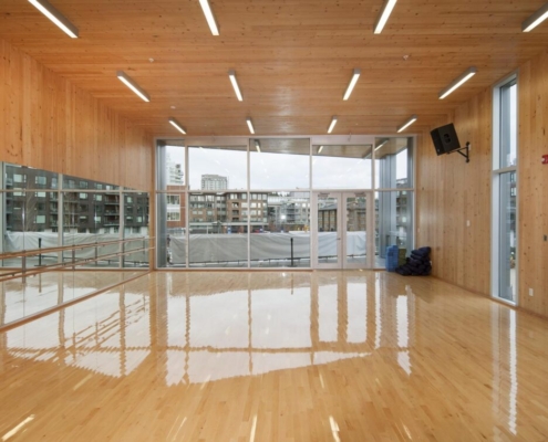 A spacious, modern dance studio with polished wooden floors, floor-to-ceiling windows, mirrored walls, and bright overhead lights. Urban buildings and a cloudy sky are visible through the windows.