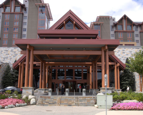 The image shows the entrance of a large, modern hotel or resort with a peaked roof, wooden beams, stone accents, and landscaped surroundings. Lush greenery and flowers frame the walkway leading to the glass doors.