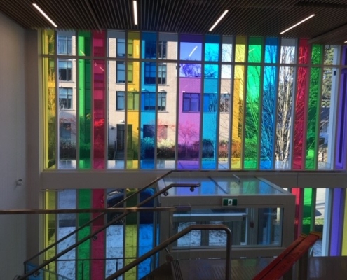 Large windows with vertical panels of colored glass in yellow, green, blue, pink, and purple. Sunlight shines through, casting colorful reflections inside; a staircase and exterior building are visible.