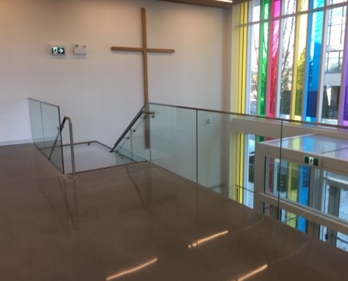 A modern interior with a large wooden cross on a white wall, glass railings, a staircase, and tall windows featuring vertical colored panels letting in natural light.