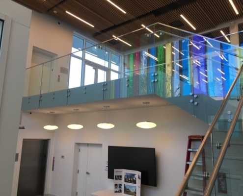 Modern interior with a glass-walled upper balcony, colorful translucent panels, suspended round lights, a TV, display board, elevator, and a ladder by a staircase. Ceiling features wooden slats and linear lighting.