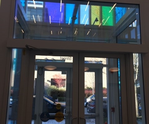 Glass double doors with a green exit sign above, colorful translucent panels overhead, and a view of parked cars and trees outside. An orange safety cone and yellow caution sticker are near the entrance.