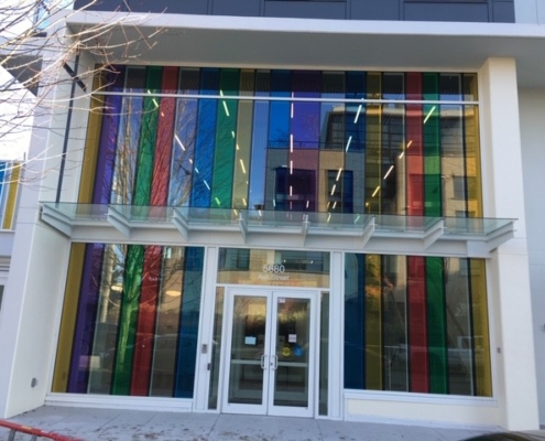 A modern building entrance with large glass doors and tall, colorful vertical panels in blue, green, red, and yellow above and beside the doors. Glass canopy covers the entrance. The address 5880 is visible above the doors.