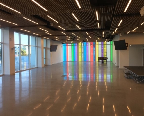 A spacious, modern room with polished floors, large windows on the left, a small stage on the right, and colorful vertical panels on the far wall. A piano is positioned near the colorful panels. Ceiling lights are lit.
