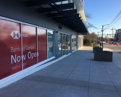 A sidewalk beside a modern building with large HSBC Banking Centre windows displaying “Now Open.” The street is quiet with few cars, planters, and some trees visible in the background.