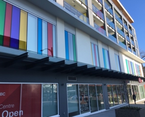 A modern building with colorful vertical window panels on the second floor, glass balconies above, and a ground-level HSBC bank with large windows and a red sign. The sky is clear and blue.