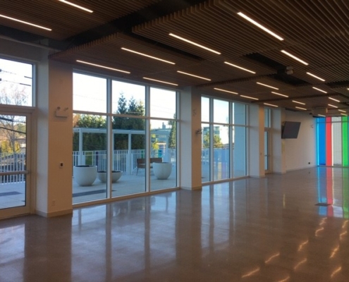 A spacious, modern room with large floor-to-ceiling windows, polished floors, a wooden slat ceiling with linear lights, and an outdoor patio visible through the windows. Colorful vertical lights are on one wall.