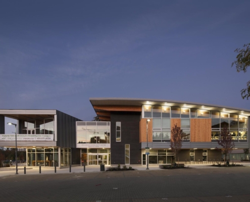 A modern, well-lit community center building with large windows and a curved roof, photographed at dusk with blue sky in the background and a few small trees in the foreground.