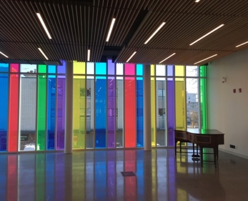 A modern room with a wooden slatted ceiling, polished floor, and large windows featuring vertical panels of colorful, translucent glass. Natural light and vivid colors fill the space; a wooden desk sits to the right.