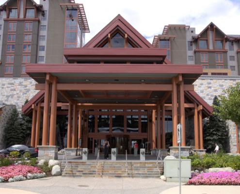 The image shows the entrance of a large hotel with a modern mountain lodge design, featuring wooden beams, stone walls, and manicured landscaping with colorful flowers and green shrubs.