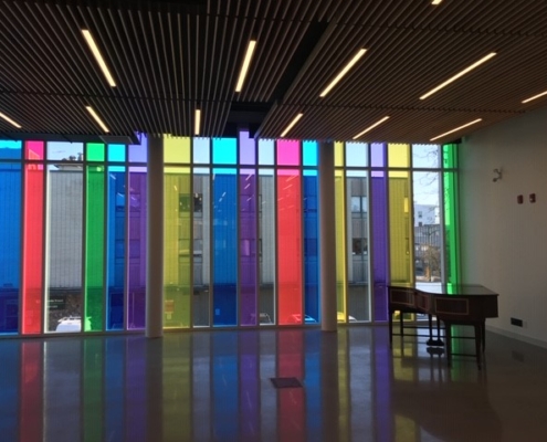 A modern interior with floor-to-ceiling windows featuring vertical panels of red, blue, yellow, green, and purple. Sunlight shines through the colorful glass, casting vibrant reflections on the shiny floor. A wooden desk sits by the wall.