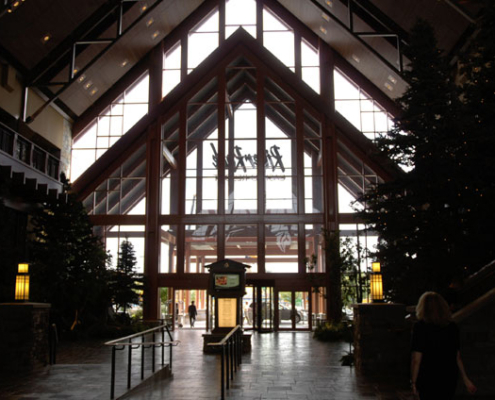 Spacious lodge interior with high, vaulted glass ceiling, large windows letting in natural light, indoor trees, soft lighting, and a few people walking toward the entrance.