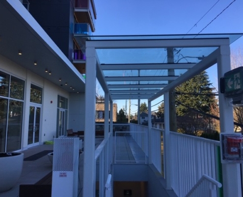 A glass-covered outdoor walkway with white railings and support beams next to a modern building. Trees and houses are visible in the background under a clear blue sky. An exit sign and fire alarm are mounted on the right.