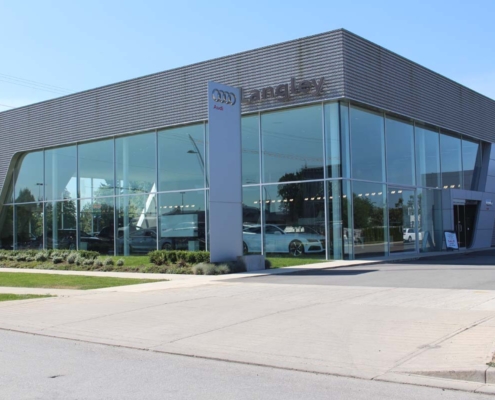 Modern Audi car dealership building with large glass windows, showcasing several vehicles inside. The exterior features sleek metal paneling and well-maintained landscaping along the sidewalk.