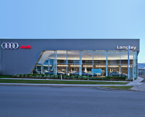 Modern Audi dealership building with large glass windows and prominent Audi and Langley signage. Several cars are visible inside the well-lit showroom, and a service center is attached on the right side of the building.