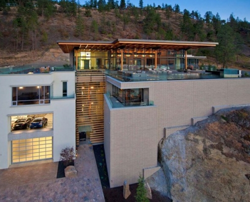 A modern multi-level house built into a hillside, featuring large windows, rooftop patios, an outdoor pool area, and a garage with two cars, surrounded by trees and rocky terrain at dusk.