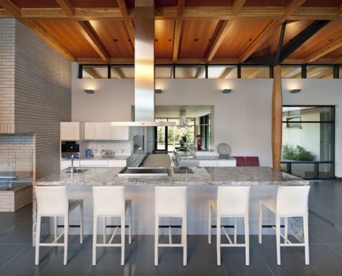 Modern kitchen with a large marble island, four white barstools, stainless steel appliances, light brick wall, wooden ceiling, and large windows letting in natural light.