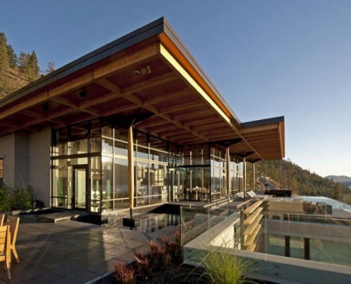 A modern house with large glass walls and a wooden roof sits on a hillside overlooking a lake, surrounded by outdoor seating and greenery, under a clear sky at sunset.