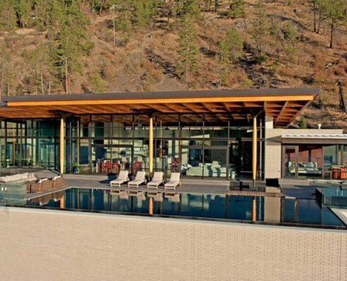 A modern luxury house with large glass windows, wooden roof beams, and an infinity pool overlooking a hillside with scattered trees. Several lounge chairs are placed by the pool.