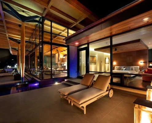 Modern luxury house at night with large glass windows, wooden ceilings, indoor and outdoor lounge areas, illuminated pool, and two cushioned chaise lounges on the patio. Warm lighting creates a cozy, inviting atmosphere.