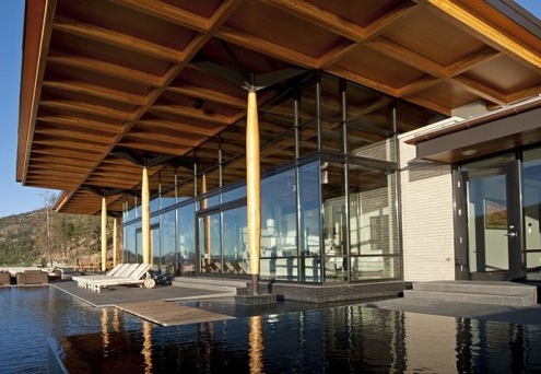 Modern house with large glass windows, wooden beams, and an overhanging roof, overlooking a reflective infinity pool and surrounded by hills under a clear blue sky. Lounge chairs are arranged by the poolside.